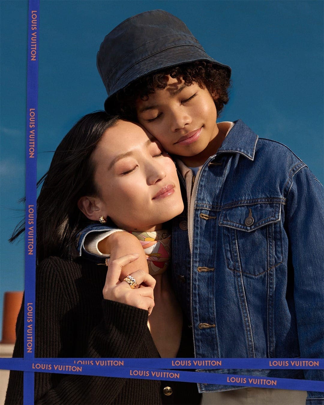 An Asian woman in a black knit top embracing a boy wearing a denim jacket and bucket hat against a blue sky background.