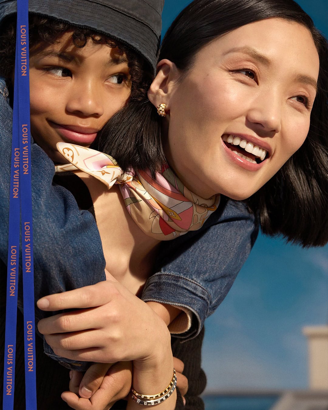 Close-up portrait of two smiling models embracing outdoors under a clear blue sky, wearing luxury accessories and branded ribbons.
