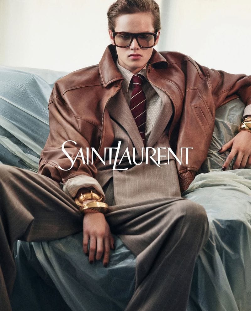 Model wearing a brown leather jacket and suit with sunglasses seated against a draped backdrop