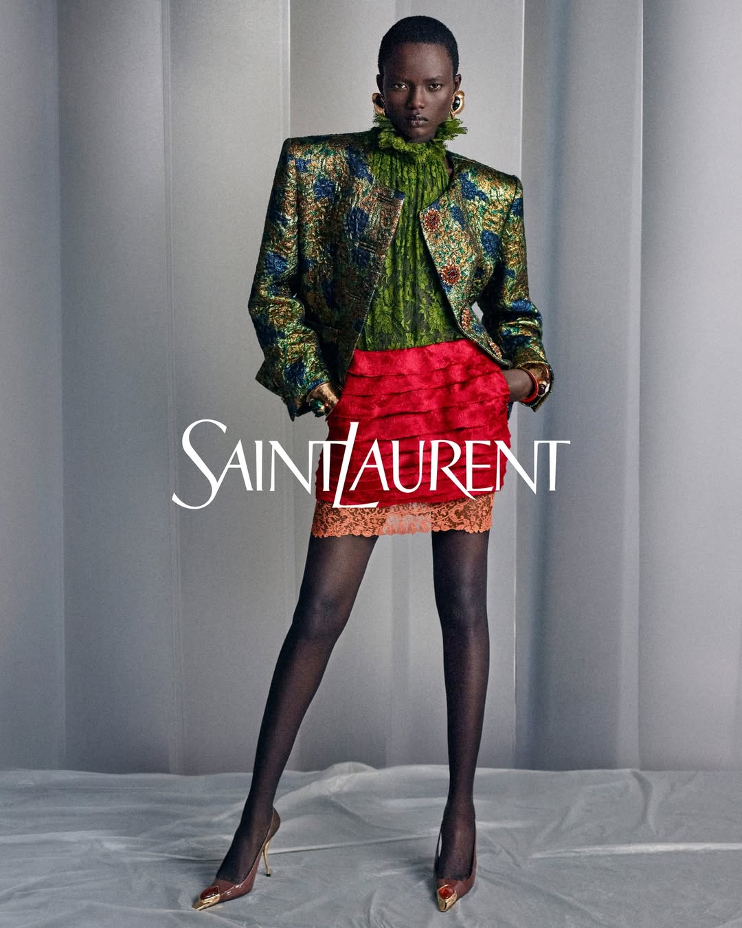 Model standing on a draped studio floor wearing a patterned blazer, green blouse, red skirt, and black tights.