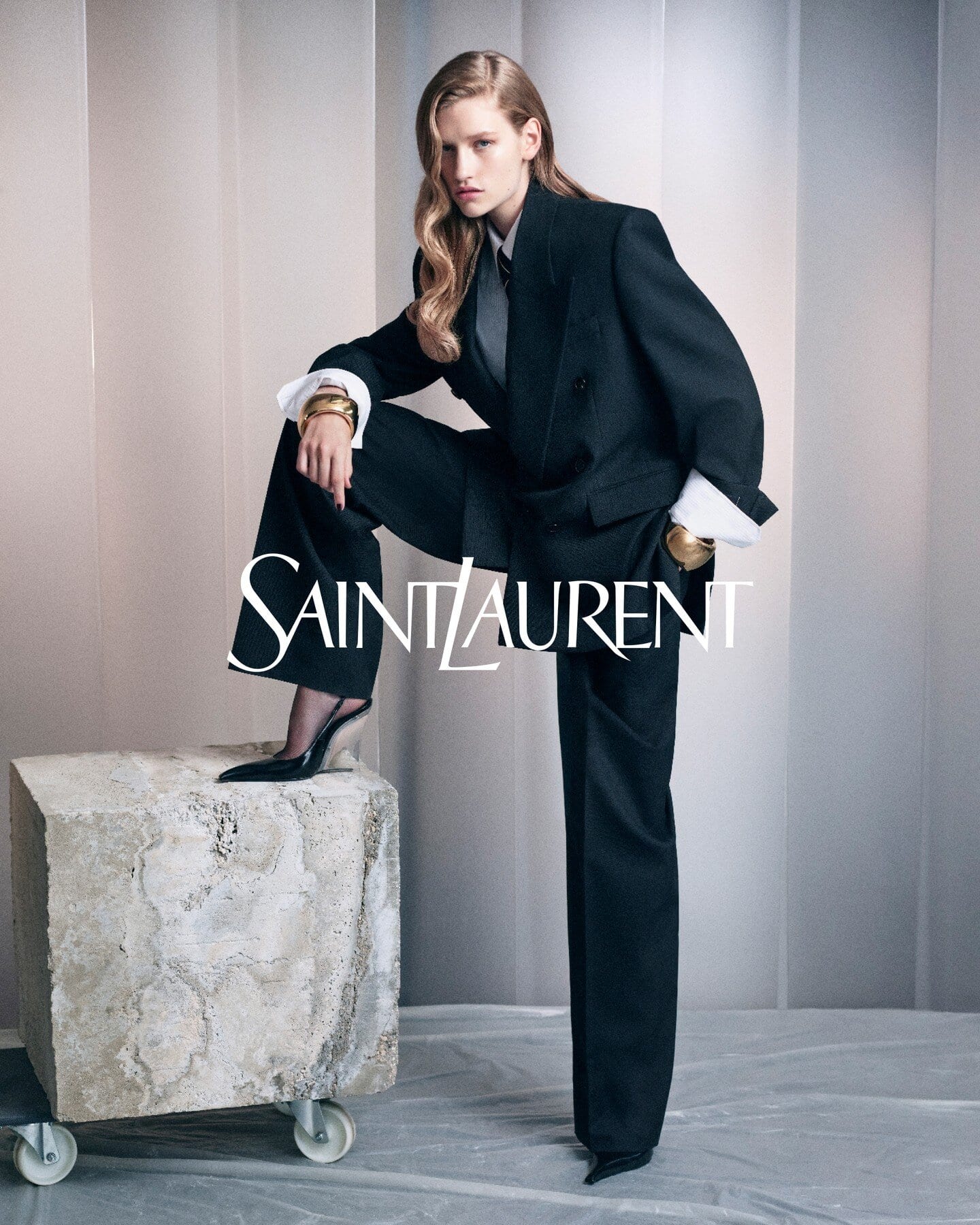 Model in black suit resting foot on a stone block with Saint Laurent text overlay against vertical panels