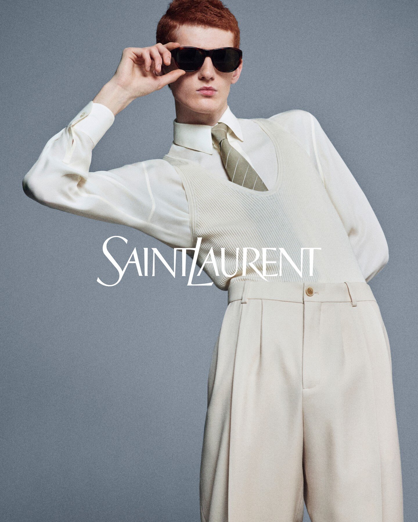 Model with red hair wearing sunglasses, white shirt, knitted vest, and tie against a grey seamless background for Saint Laurent.
