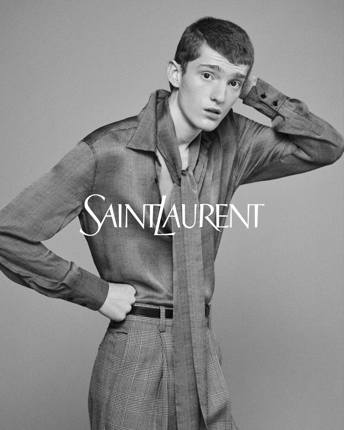 Monochrome fashion portrait of a male model wearing a patterned collared shirt and tie against a grey seamless studio backdrop.