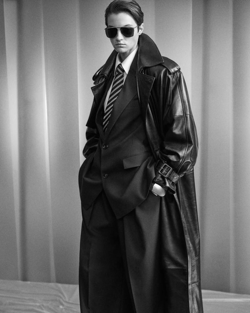 Model in dark sunglasses wearing a black leather trench coat over a tailored suit against a pleated background