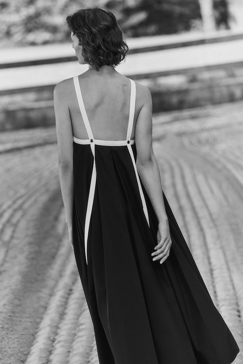 Model seen from behind wearing a long black dress with thin white shoulder straps outdoors.