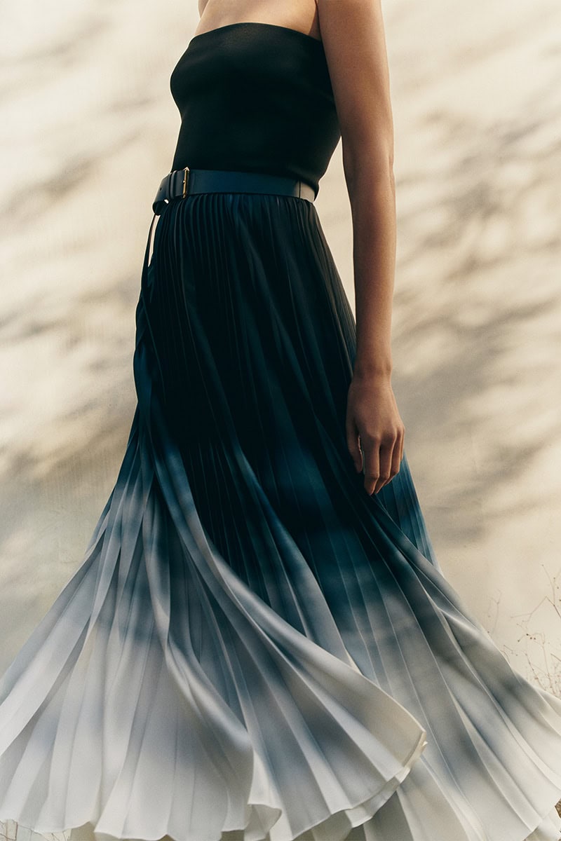 Model wearing a black top and ombré pleated skirt with a belt against a sunlit wall in the Leisure Spring 2025 campaign by Giada.