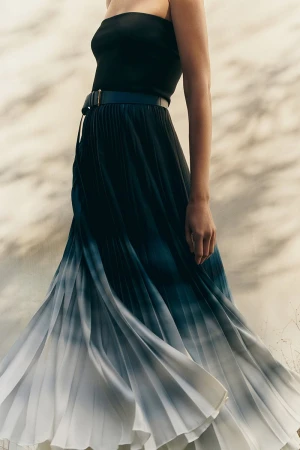Model wearing a black top and ombré pleated skirt with a belt against a sunlit wall in the Leisure Spring 2025 campaign by Giada.