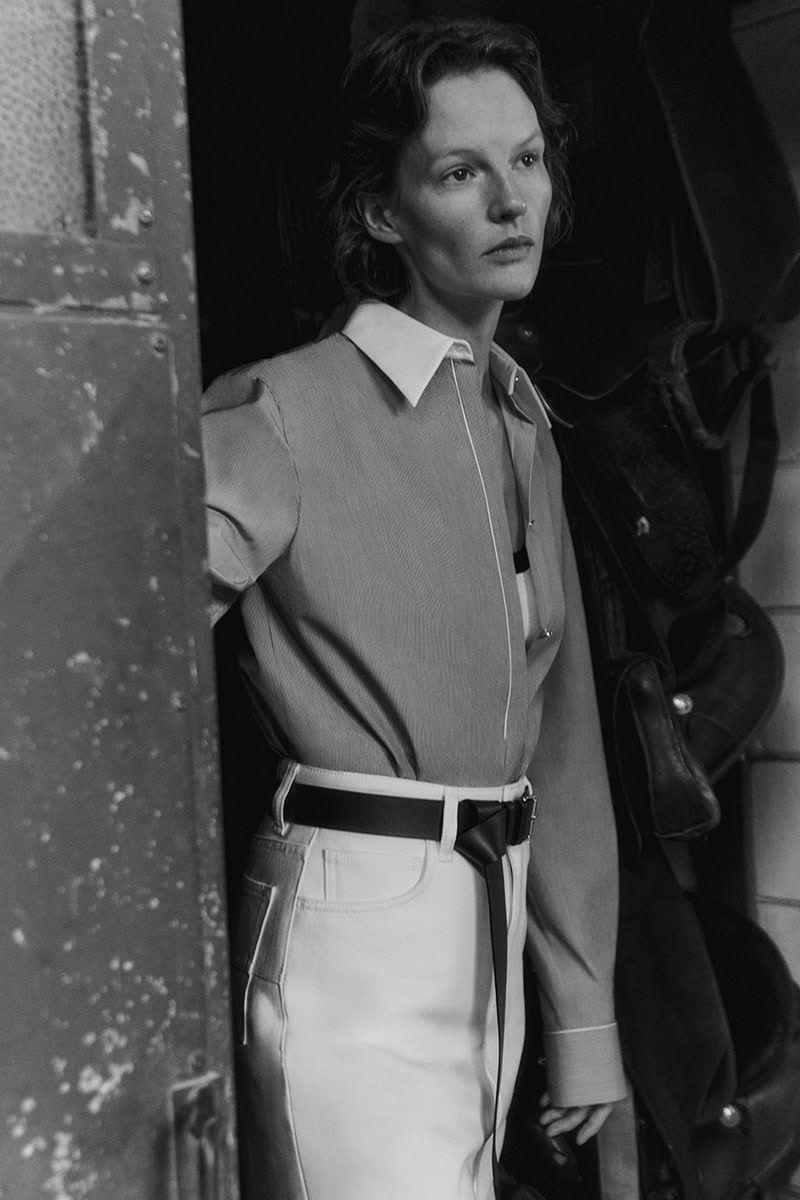 Black and white portrait of a female model wearing a button-down shirt and belted trousers in an interior setting.