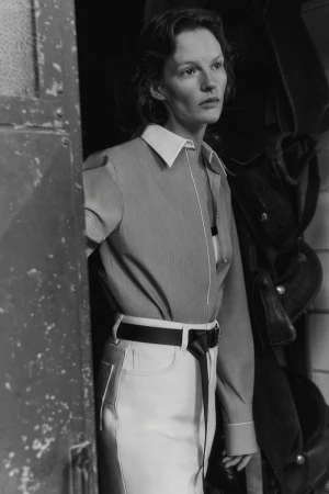 Black and white portrait of a female model wearing a button-down shirt and belted trousers in an interior setting.