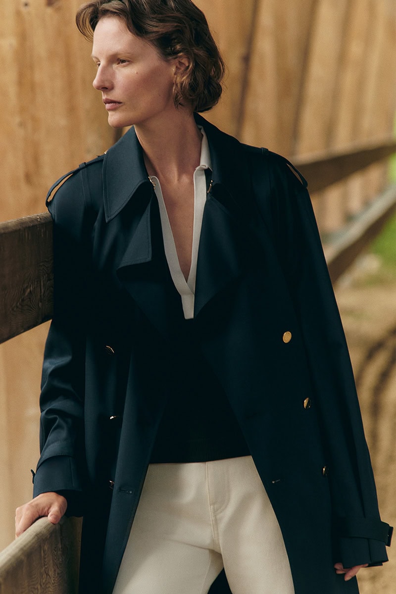 Model Sara Blomqvist in a dark trench coat leaning against a wooden fence for Giada Leisure campaign