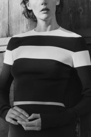 Model wearing a black and white striped long-sleeve top against a dark wooden plank wall in a black and white portrait.