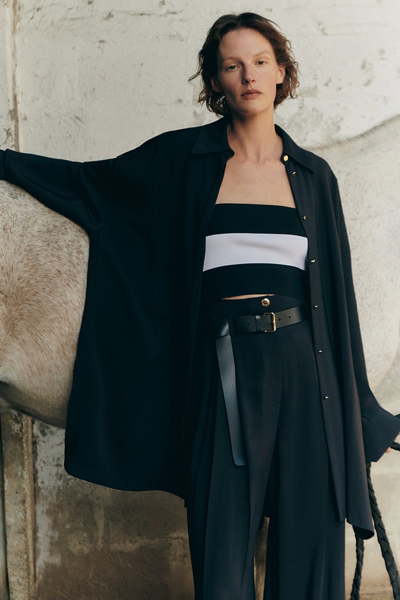 Model wearing a black shirt and striped bandeau posing next to a white horse against a textured rustic wall.