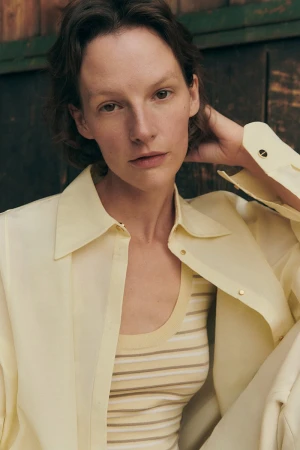 Model Sara Blomqvist wearing a pale yellow button-down shirt and striped top against a dark wood background
