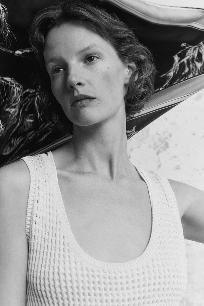 Model with short wavy hair wearing a white textured tank top against a textured wall with fabric draped behind