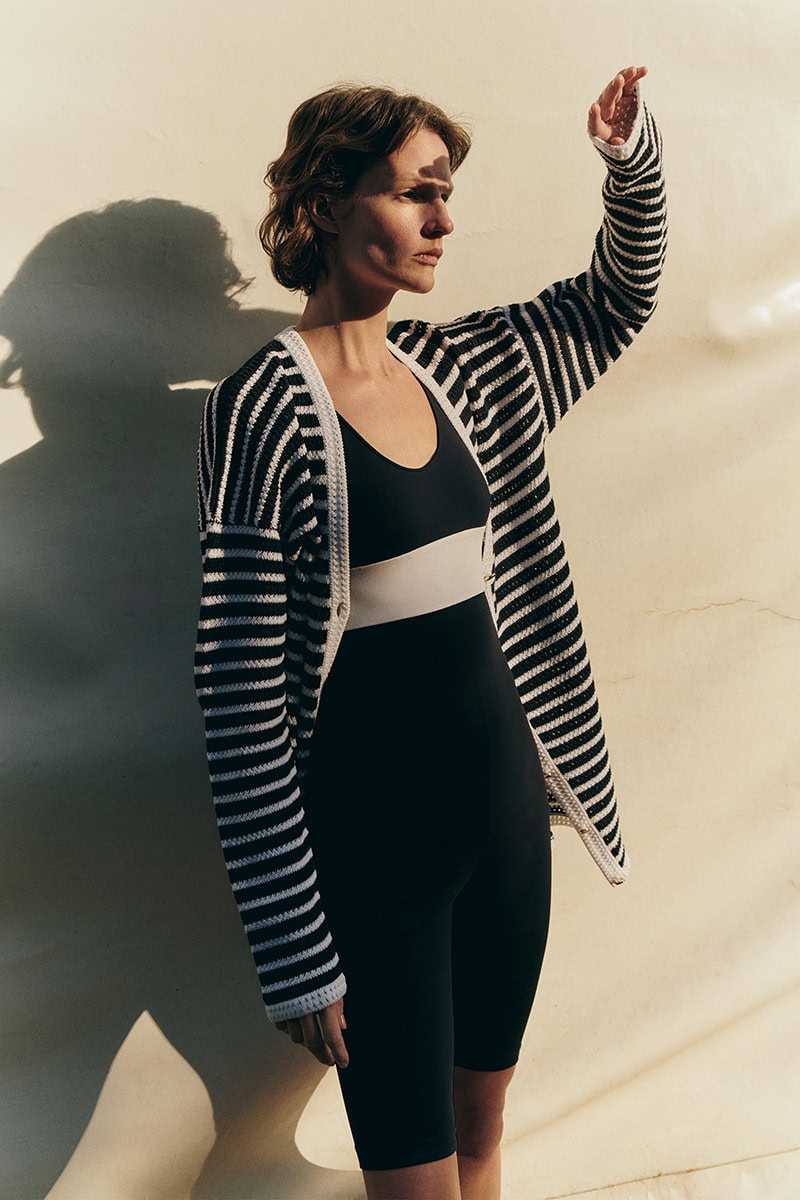 Model wearing a black jumpsuit and striped cardigan raising her arm against a pale wall with dramatic light and shadow.