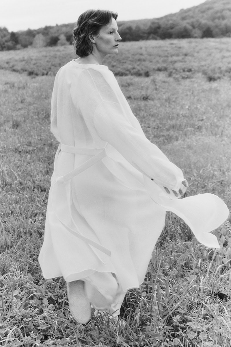 Monochrome fashion campaign image of Sara Blomqvist walking through a grassy meadow wearing a light belted coat by Giada.