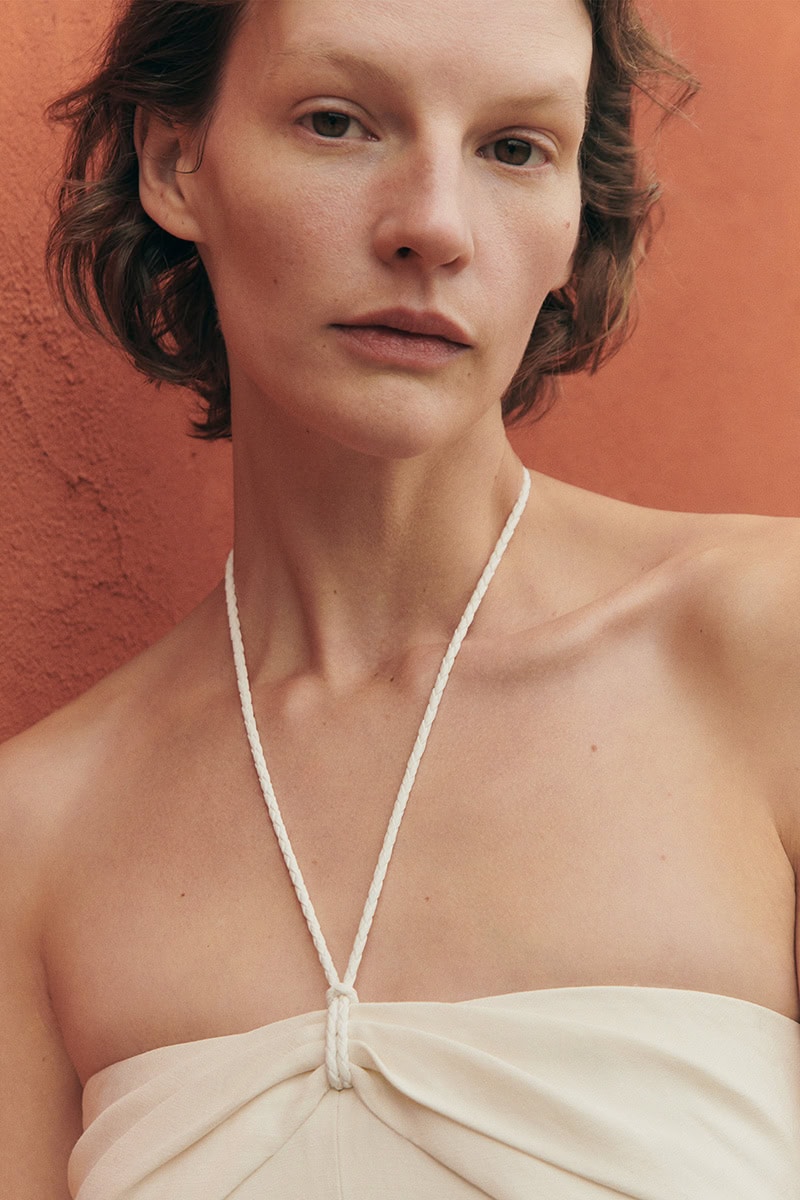 Close-up portrait of a female model wearing a white top with a halter neck string against a terracotta-colored wall.