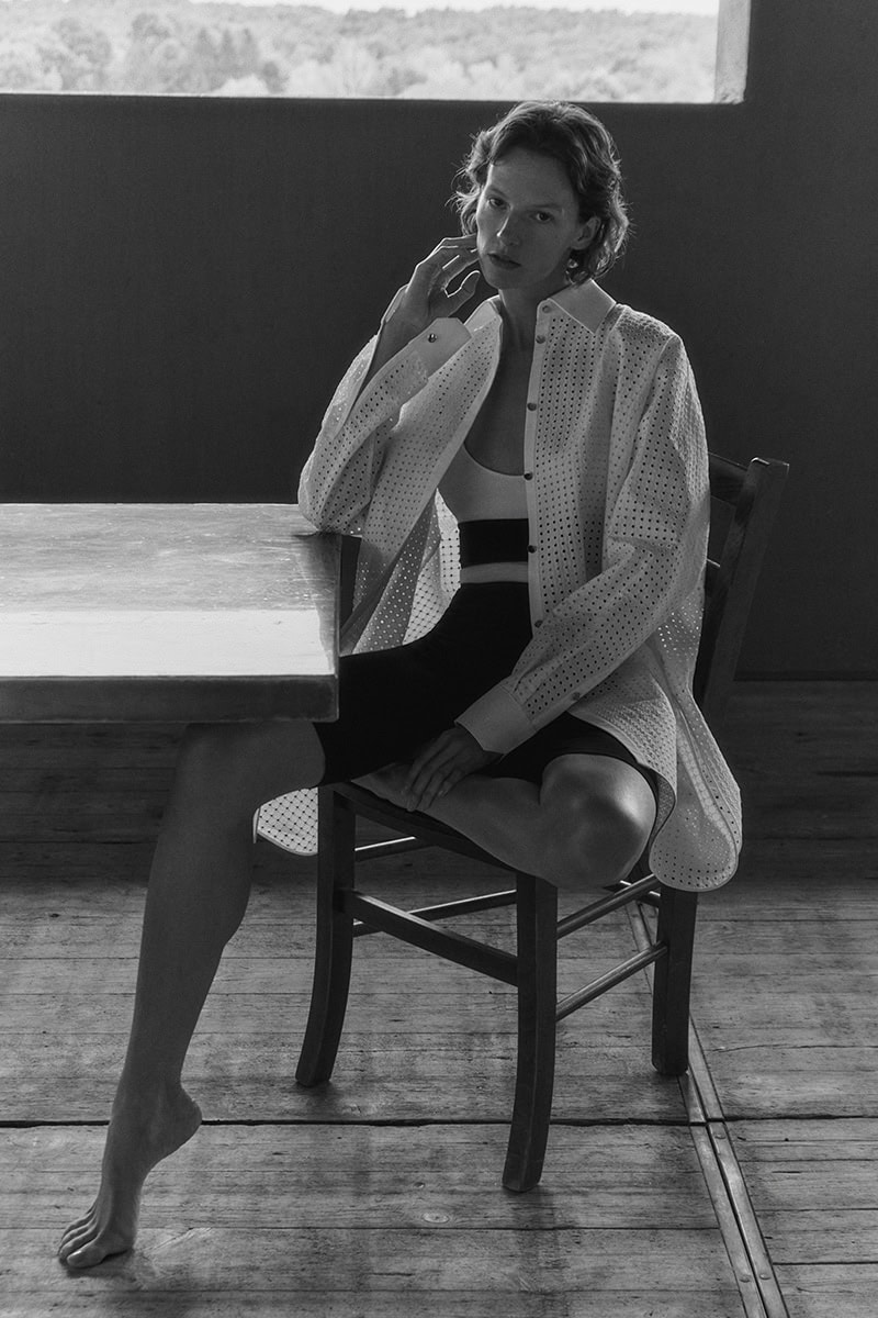 Model sitting on a wooden chair next to a table wearing an unbuttoned white patterned shirt and black shorts