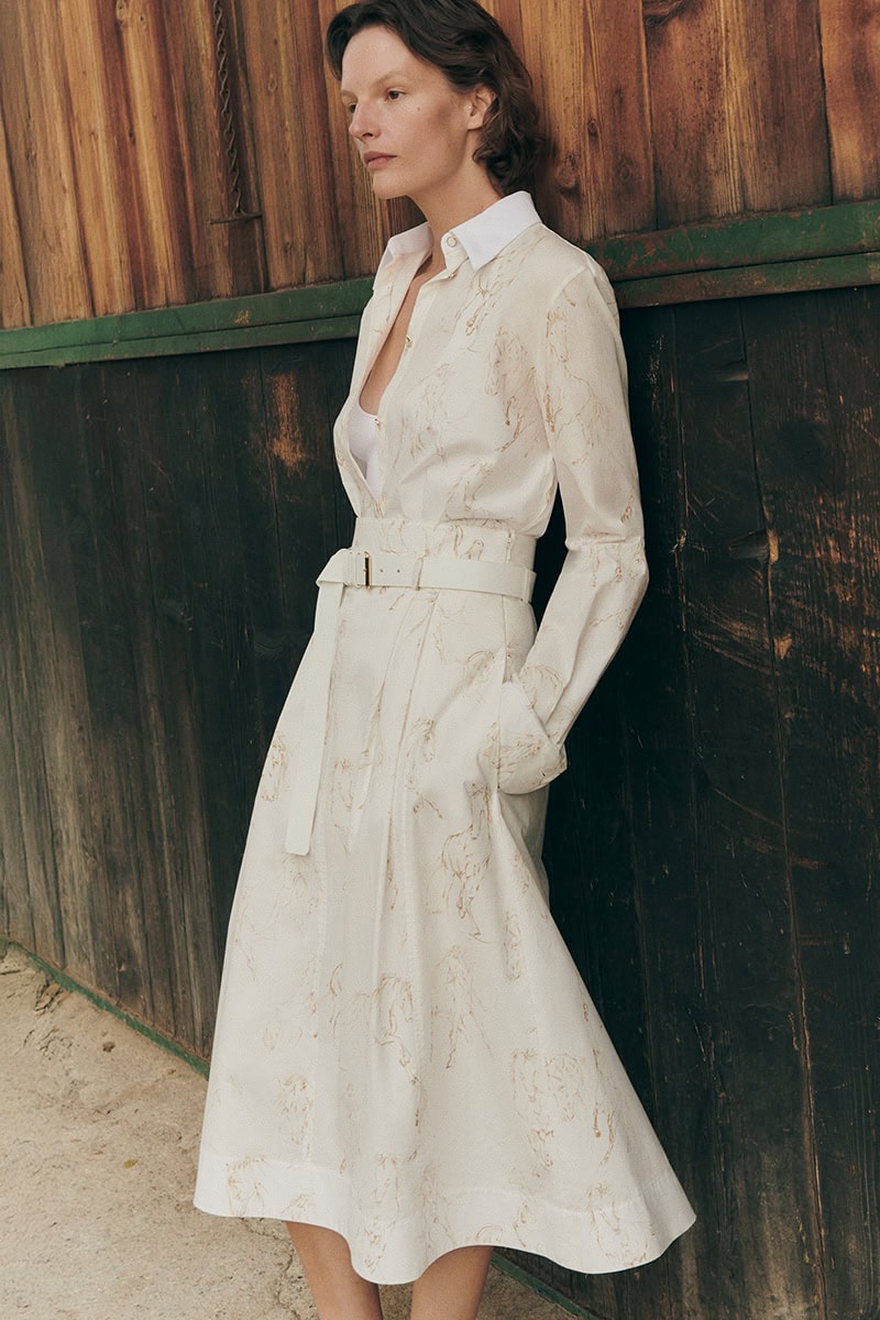 Model leaning against a rustic wooden wall wearing a white patterned belted dress and long sleeve blouse.
