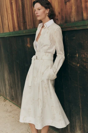 Model leaning against a rustic wooden wall wearing a white patterned belted dress and long sleeve blouse.