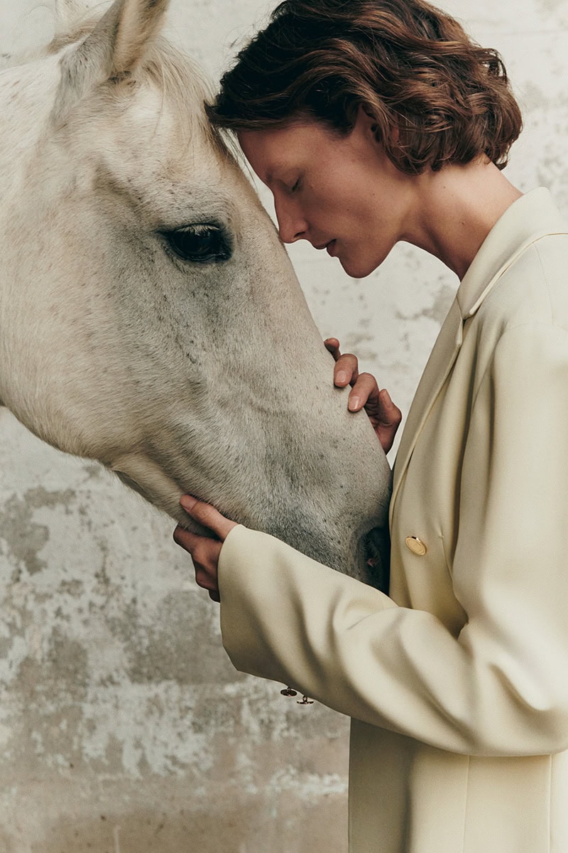 Model Sara Blomqvist in a beige blazer gently touching a white horse head against a textured neutral background