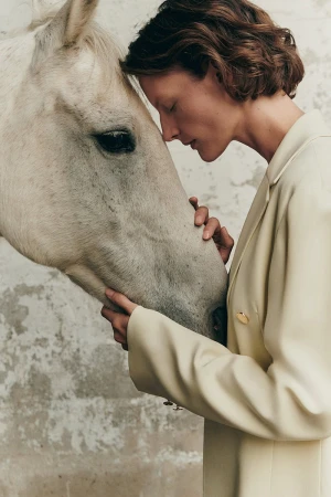 Model Sara Blomqvist in a beige blazer gently touching a white horse head against a textured neutral background
