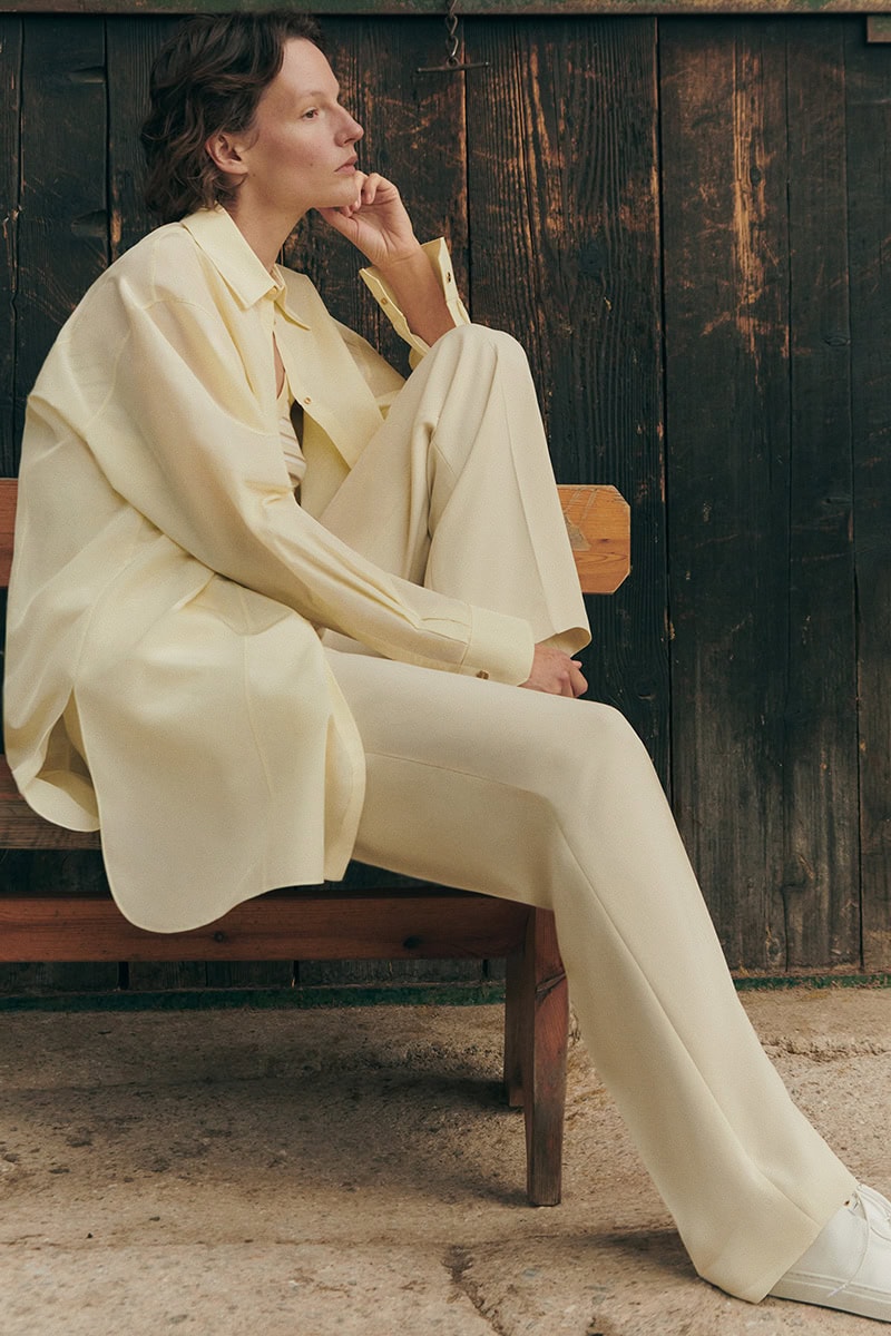 Model wearing a light yellow matching shirt and trousers seated on a wooden bench outdoors against a dark wood wall.