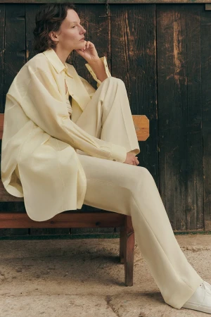 Model wearing a light yellow matching shirt and trousers seated on a wooden bench outdoors against a dark wood wall.