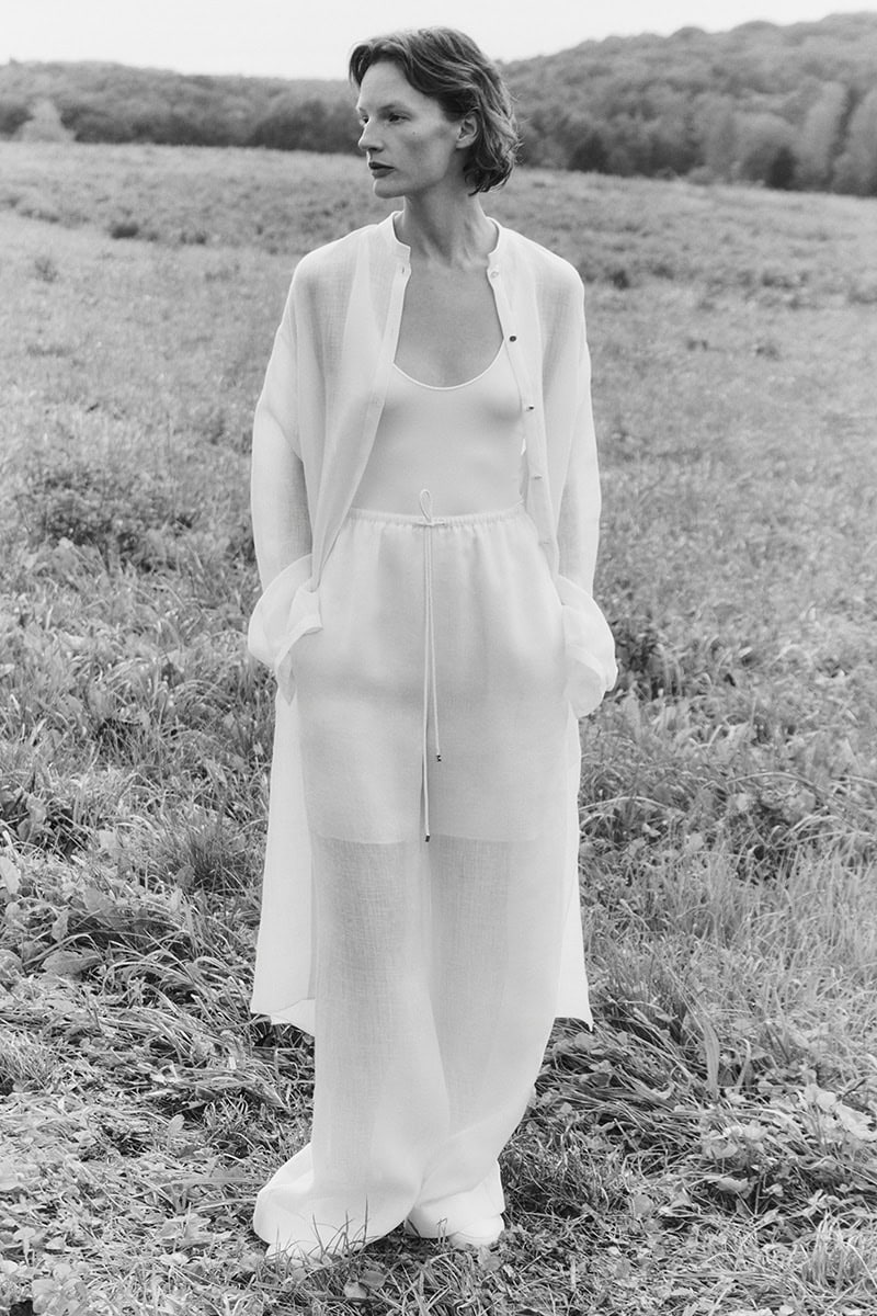 Monochrome fashion campaign image of a female model standing in a grassy field wearing light trousers and a cardigan