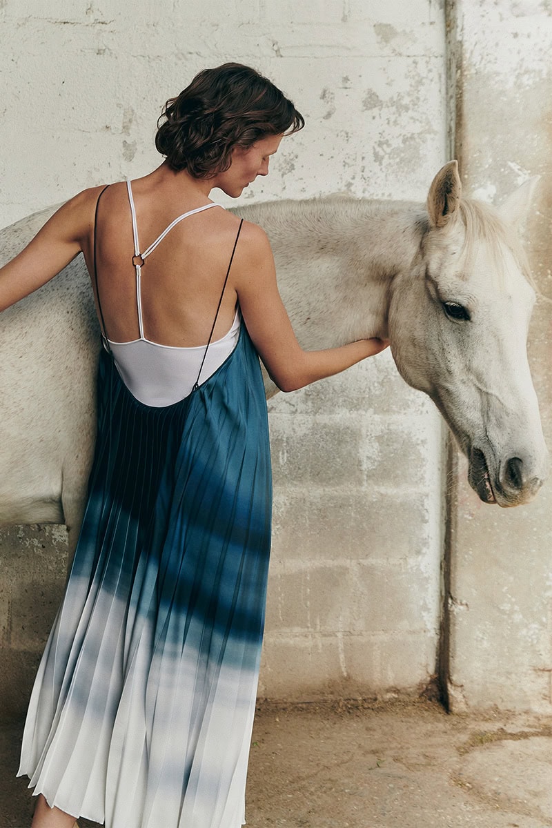 Model wearing a blue ombre pleated dress standing beside a white horse in front of a textured stone wall