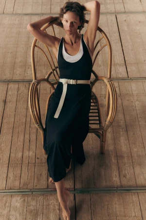 Model wearing a dark dress with white belt sitting in a wicker chair on a wooden deck