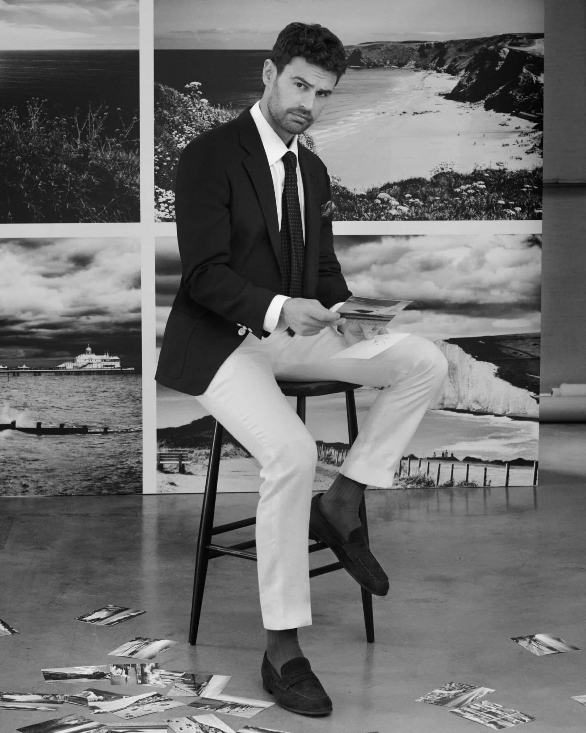 A man in a black suit jacket and white trousers sitting on a stool holding photographs in a studio with a scenic photo backdrop.