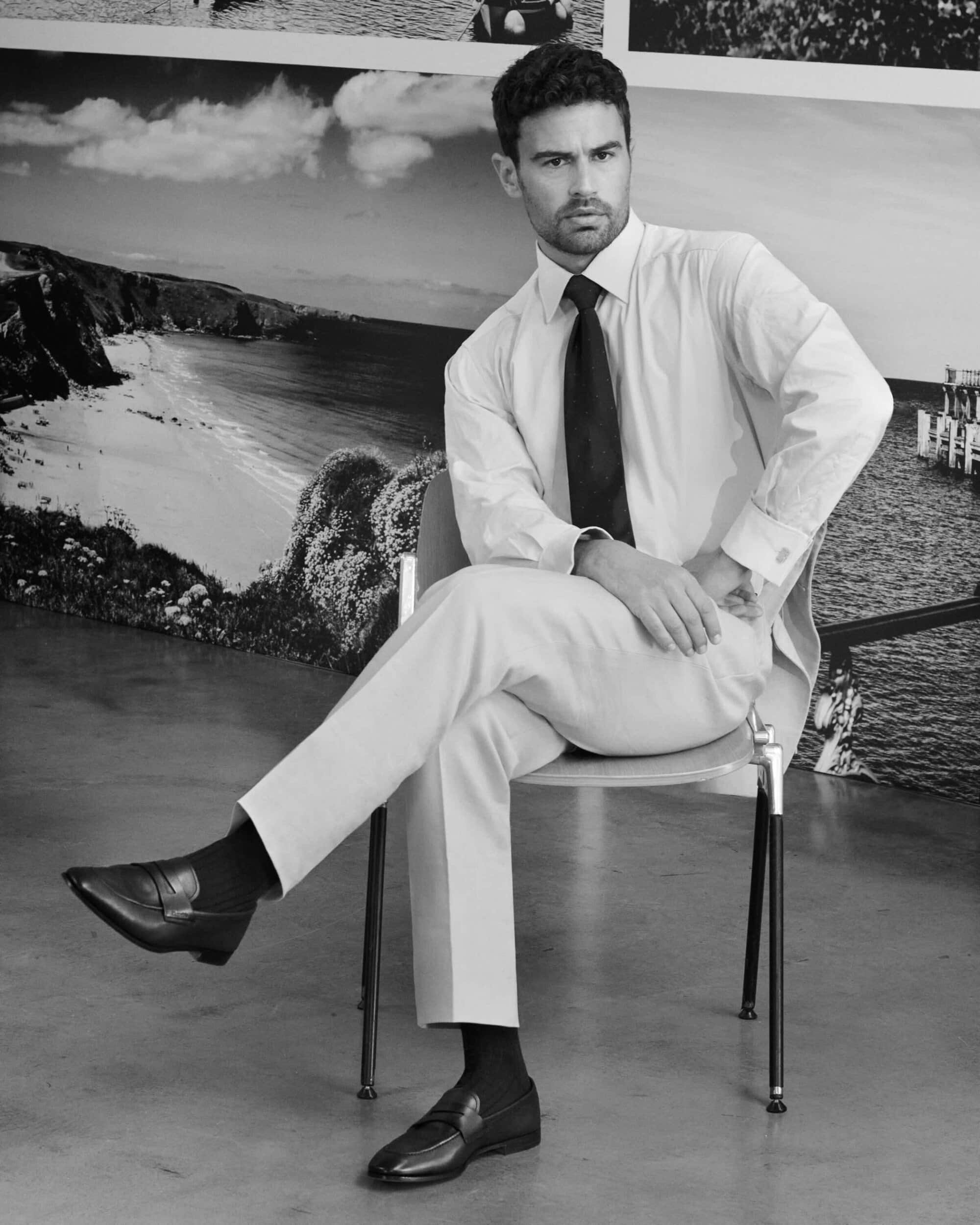 Monochrome portrait of a man seated on a chair wearing a button-down shirt, tie, trousers, and leather loafers against a scenic backdrop.