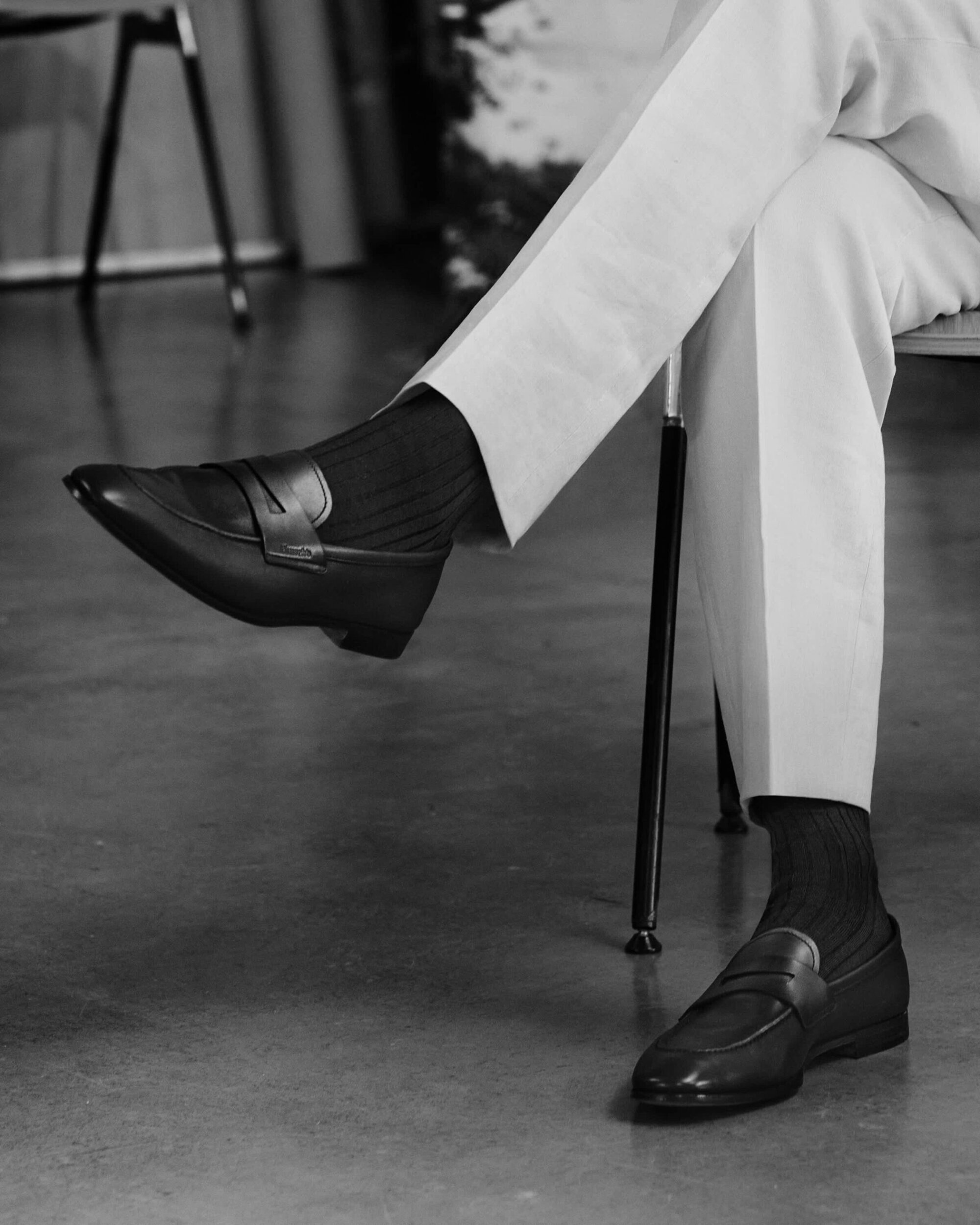 Close-up of a person sitting with crossed legs wearing dark leather loafers and ribbed socks with light trousers.