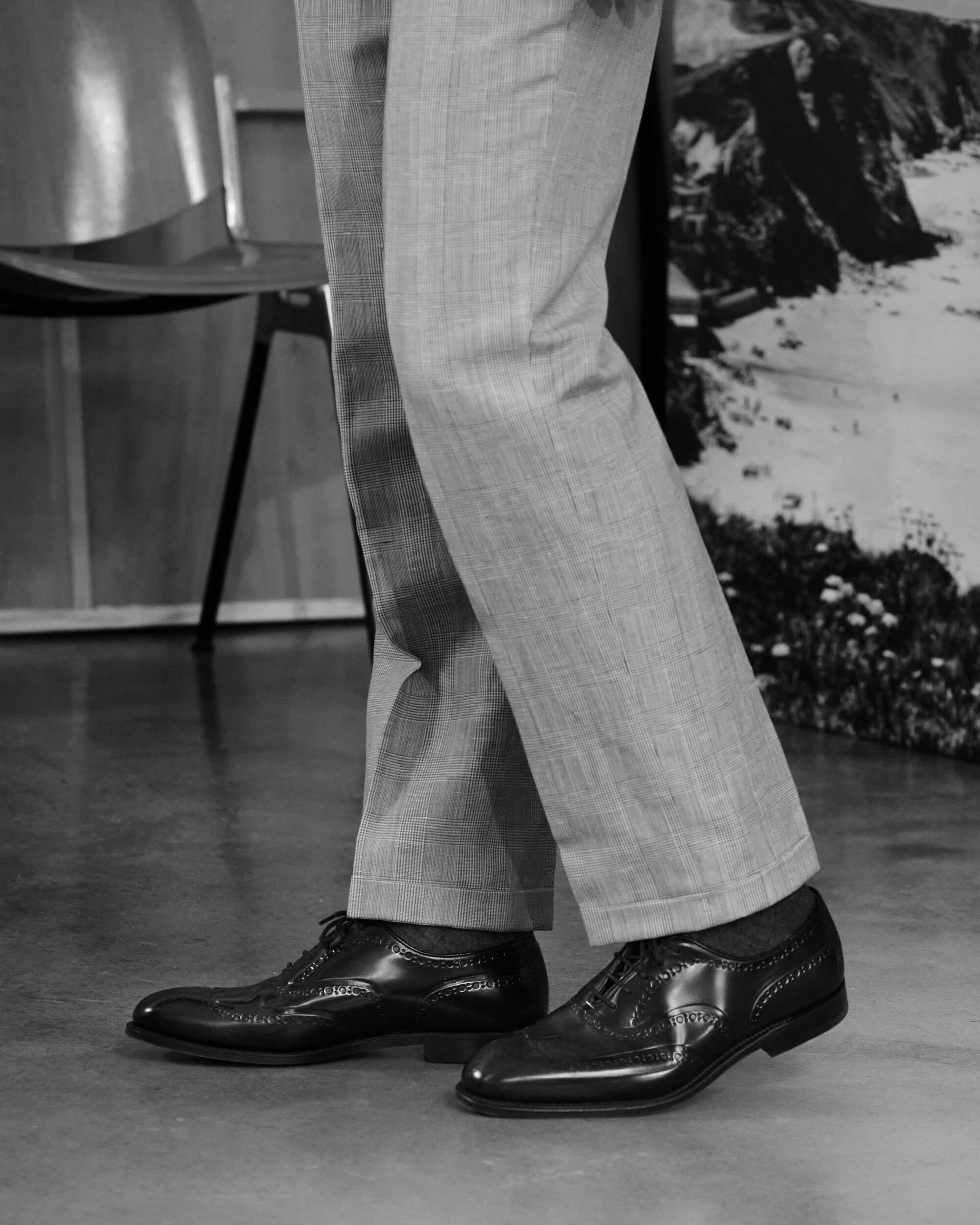 Close-up shot of a person wearing light trousers and polished black leather brogues against a studio background.