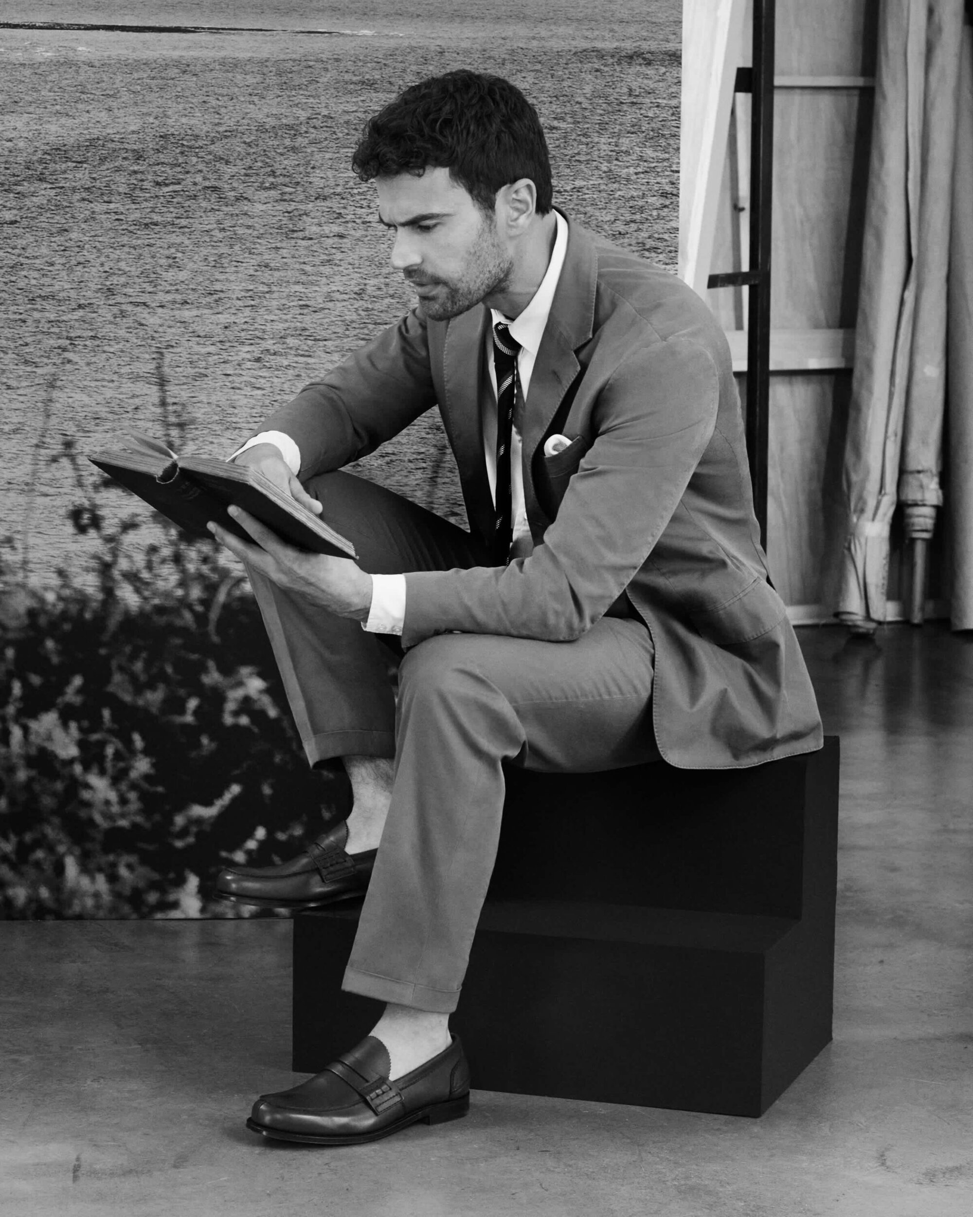 Man in suit and loafers seated on a block reading a book in a studio with a scenic backdrop for Church's Stage Craft campaign