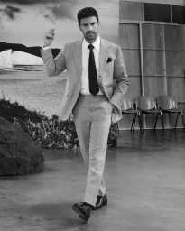 Monochrome portrait of Theo James wearing a tailored light suit and dark tie against a studio set backdrop with a landscape print