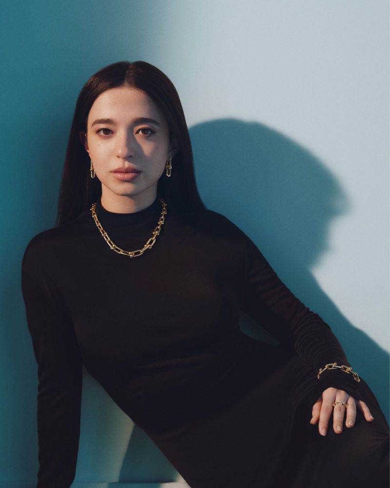 Woman in black turtleneck wearing gold link necklace and matching earrings against a blue wall