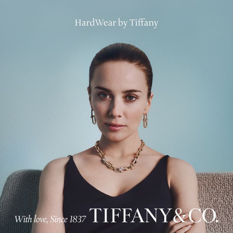 Portrait of a woman wearing gold chain jewelry and a dark tank top against a soft blue background for Tiffany and Co.