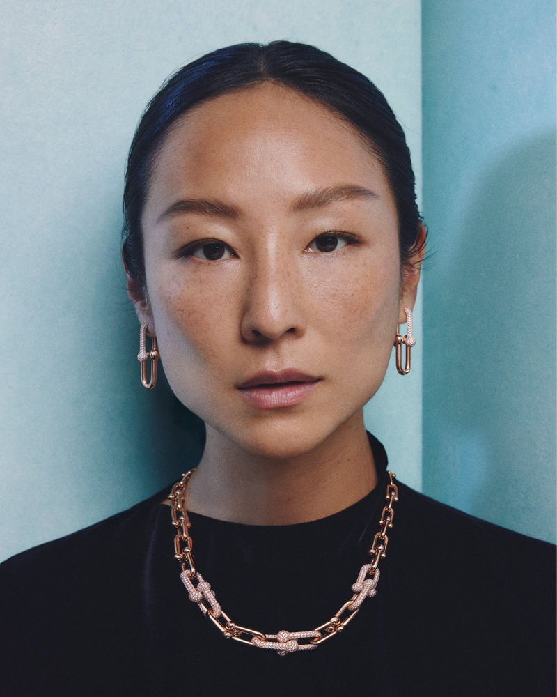 Portrait of a woman wearing a gold link necklace and matching link earrings against a light blue corner wall.