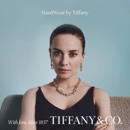 Portrait of a woman wearing gold chain jewelry and a dark tank top against a soft blue background for Tiffany and Co.
