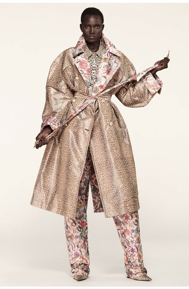 A tall, dark-skinned woman models a tan animal-print trench coat with a floral-print lining over matching floral pants on a plain background.