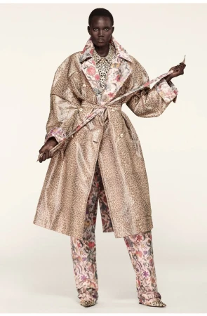 A tall, dark-skinned woman models a tan animal-print trench coat with a floral-print lining over matching floral pants on a plain background.