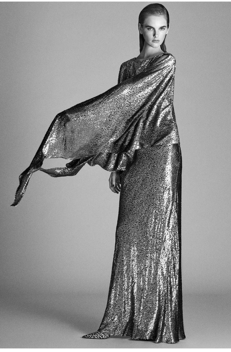 Elegant fashion shot of a solitary, slender adult woman in a floor-length, sparkling silver gown with wide, flowing sleeves against gray backdrop.