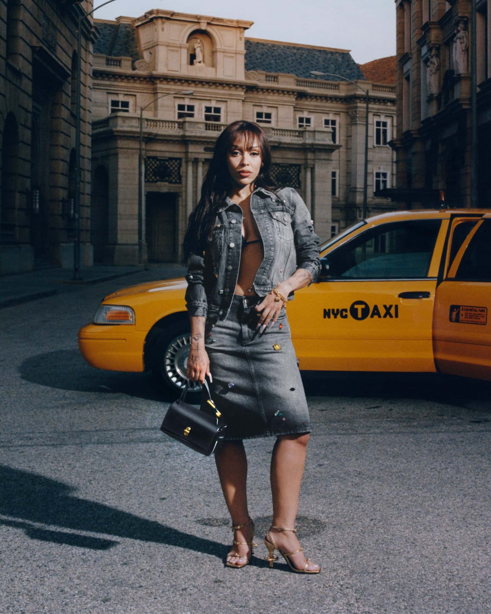 Doja Cat posing in a grey denim jacket and skirt holding a handbag in front of a yellow NYC taxi cab