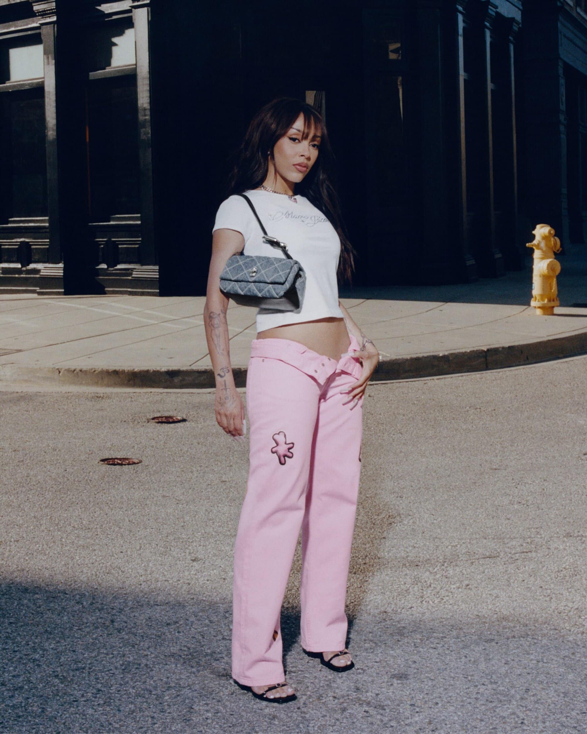 Doja Cat wearing a white t-shirt and pink pants with a quilted handbag standing on a city street.