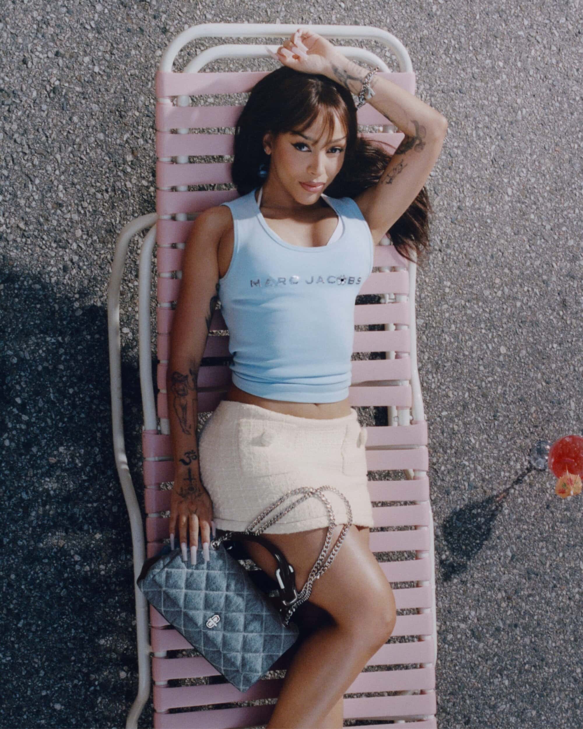 Doja Cat lounging on a pink chair wearing a blue tank top and tweed skirt for Marc Jacobs Staycation campaign