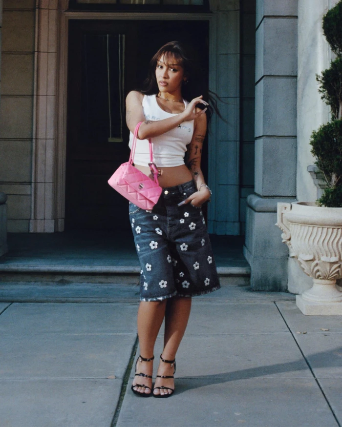 Doja Cat wearing a white tank top and floral denim shorts holding a pink handbag outside a building for Marc Jacobs