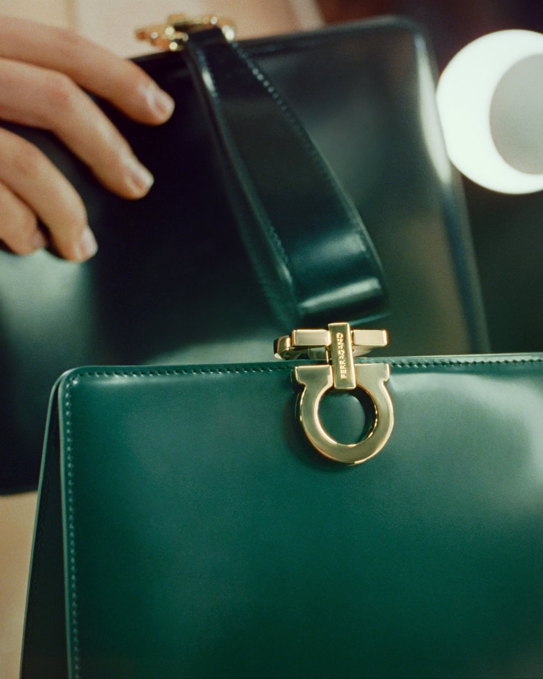 Close-up of glossy green leather Ferragamo handbags with gold hardware held by a person.