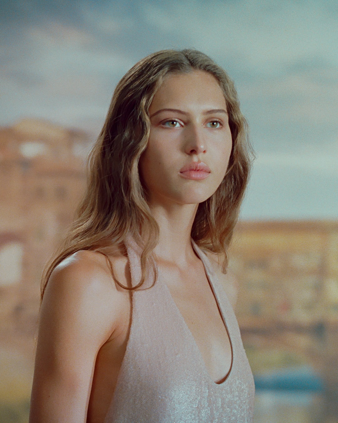 Portrait of a female model wearing a pale halter dress against an atmospheric painted backdrop with distant architecture.